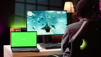 Mockup laptop next to man playing high FPS singleplayer videogame with spaceship shooting lasers. Isolated screen notebook near gamer using crosshair to hit targets in SF game video