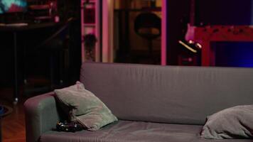 Man arriving home, gaming and enjoying leisure time. Man sitting on couch in neon illuminated living room, playing videogame on console, relaxing after hard day at work, camera A video
