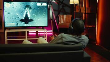 Man playing entertaining spaceship flying videogame on large smart TV, wearing headphones. Player enjoying SF arcade game on gaming console, piloting spacecraft in galaxy using gamepad, camera B video