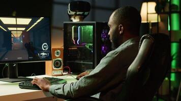 Smiling man playing games on gaming PC at computer desk, enjoying day off from work. Upbeat gamer battling enemies in online multiplayer shooter from neon lights ornate home, camera A video