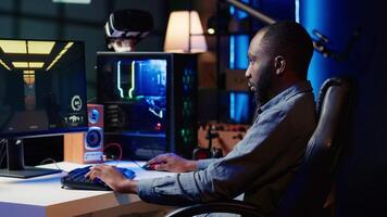Portrait of smiling man playing FPS videogame on PC screen in neon illuminated apartment living room. Cheerful gamer enjoying shooter game on computer desktop display in RGB lit home, camera A video