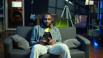Cheerful man wearing headphones, celebrating after victory in videogame, neon lit apartment. Person on couch throws fists in air from excitement after winning game on console with controller, camera B video