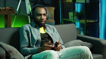Cheerful man wearing headphones, celebrating after victory in videogame, neon lit apartment. Person on couch throws fists in air from excitement after winning game on console with controller, camera A video