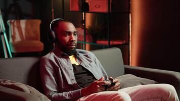 Tired gamer in RGB lit home late at night gaming with gamepad. Man sitting on sofa in neon illuminated apartment, playing videogames on console using controller, looking exhausted, camera A video