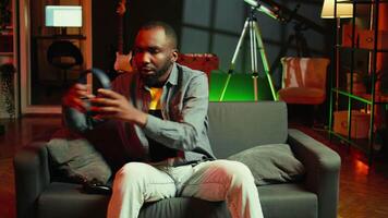 Player in RGB lit home putting headphones on to be immersed in videogame experience, enjoying relaxation time. Man wearing headset sitting on couch, playing games on console using gamepad, camera A video