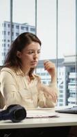 Vertical Young entrepreneur listening to a podcast while she solves daily business tasks in high end office, corporate success story. Female director working with headphones, determination. Camera B. video