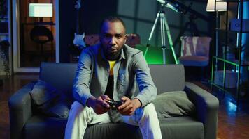 Player in RGB lit home gaming with joystick and enjoying relaxation time. Man sitting on sofa in neon illuminated living room, playing videogames on console using controller, camera B video