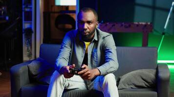 Player in RGB lit home gaming with joystick and enjoying relaxation time. Man sitting on sofa in neon illuminated living room, playing videogames on console using controller, camera A video