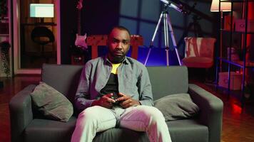Focused gamer in RGB lit home gaming with gamepad, enjoying himself. Man sitting on sofa in neon illuminated living room, playing videogames on console using controller, camera B video