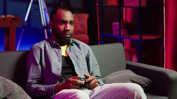 Focused gamer in RGB lit home gaming with gamepad, enjoying himself. Man sitting on sofa in neon illuminated living room, playing videogames on console using controller, camera A video