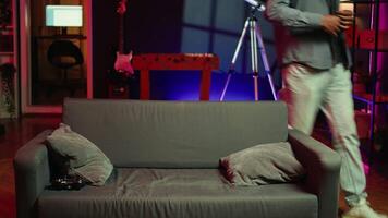 Man arriving home, gaming and enjoying leisure time. Man sitting on couch in neon illuminated living room, playing videogame on console, relaxing after hard day at work, camera B video