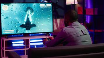 Gamer using controller and headphones to play first person shooter videogame on large smart TV. Man in home theatre using high tech gaming console gamepad to defeat foes, camera A video