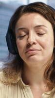 Vertical Female entrepreneur enjoys multitasking with her favorite music on headset, singing and dancing at her CEO desk. Woman with leadership skills working on her business with songs. Camera A. video