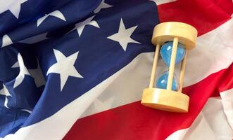 Hourglass on table with USA flag in the background. Sand glass on old wooden table with American flag in the background, vertical image. photo