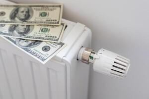 Stack of dollar banknotes on heating radiator battery with thermostat temperature regulator. Concept of expensive heating costs and rising energy bill prices for winter cold season. photo