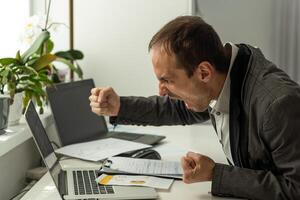 Angry annoyed frustrated caucasian business man, sitting at workplace with laptop, screaming, gesturing with fists, failed deal, disappointed with project result, experiencing negative emotions photo