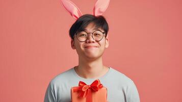 Photo of cheerful, positive Asian man with rabbit ears holding easter gift isolated on pastel color background with copy space
