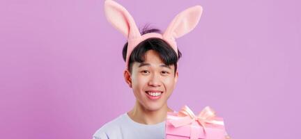 Photo of cheerful, positive Asian man with rabbit ears holding easter gift isolated on pastel color background with copy space