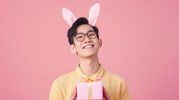 Photo of cheerful, positive Asian man with rabbit ears holding easter gift isolated on pastel color background with copy space