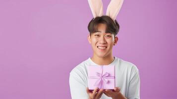 Photo of cheerful, positive Asian man with rabbit ears holding easter gift isolated on pastel color background with copy space