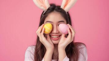 Happy beautiful young woman smiling wearing rabbit ears holding colorful Easter eggs front eyes, Asian female with bunny ear hold easter egg covering eye, studio shot isolated on pink background photo