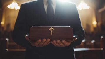 Priest in black suit standing in church and carefully holding wooden box with golden cross on it photo