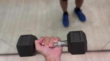 hand gripping dumbbell with a strong, determined hold. The background features a blurred view of other gym equipment, highlighting the focus on the dumbbell and the intensity of the workout, fitness video