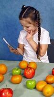 Little girl holding tablet in the kitchen. Sweet fruits laying on the table. Cute child playing games on gadget near the table with fresh apples and oranges. Vertical video