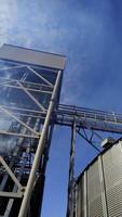 Modern elevator complex for agriculture. Metal granary against blue sky. Smoke released in the air from industrial equipment for grain processing. View from below. Vertical video