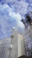Modern plant with metal elevator for agribusiness. White smoke from industrial grain dryer on sky background. Storage and further processing of grain. View from below. Vertical video