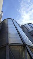 Modern granary under blue sky. Large aluminum containers for storing grains. Exterior of grain elevators. Close-up. View from below. Vertical video