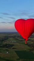 Flight of a red airship. Beautiful hot air balloon in heart shape travelling over the green fields in the morning. Aerostat with basket in the sky. Vertical video