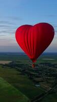 rosso dirigibile nel il forma di un' cuore. caldo aria Palloncino nel il cielo al di sopra di un' campo nel il campagna volante nel il bellissimo sera cielo. verticale video