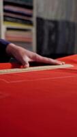 Woman tailor is working at table with red cloth in atelier. Skilled seamstress is drawing sketch lines with a soap or a chalk and a ruler on red fabric. Vertical video