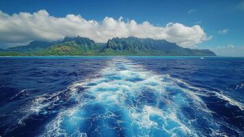 Vibrant ocean waves trail behind a boat near lush green mountains under a clear blue sky photo