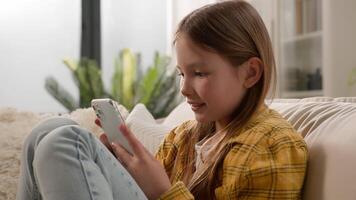 Smiling carefree little Caucasian kid girl scrolling typing screen playing games online using mobile phone telephone application. Child daughter schoolgirl browsing Internet couch home. Device overuse video
