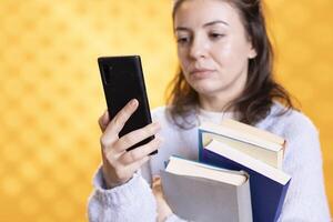 Woman typing chat messages on cellphone while holding stack of books, isolated over studio background. Student with pile of academic textbooks in arms texting mates on phone, close up shot photo