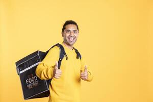 Portrait of confident man holding thermal backpack, doing thumbs up sign, studio background. Radiant delivery person prepared to deliver takeaway food to customers, doing positive hand gesture photo