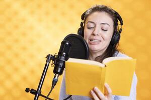 Smiling voice talent reading book, recording audiobook using microphone and headset, studio background. Joyous lady using mic and headphones to produce digital recording of novel photo