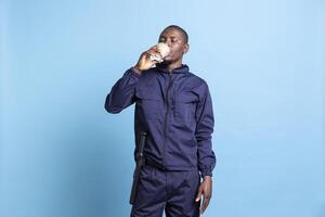 Portrait of african american security guard serves coffee cup in studio, enjoying a caffeine beverage against blue background. Patrol employee with uniform and baton drinking refreshment. photo