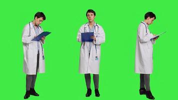Physician taking notes after patient examination against greenscreen backdrop, writing down medicaments to cure illness. Doctor in white coat using clipboard papers for checkup. Camera A. photo