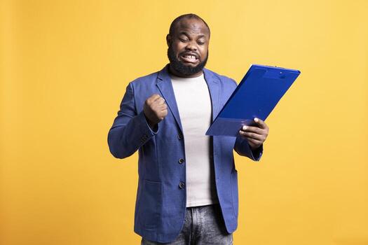 Man celebrating while browsing though financial report pages, happy with good job, studio background. Joyous worker exalting while reading paperwork, satisfied with good profits turnover photo