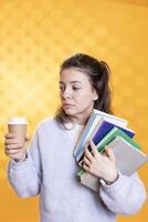 Nervous woman with troubled look on face carrying books and coffee cup, studio background. Student feels disturbed before university assessment, holding textbooks, drinking caffeine to gain courage photo