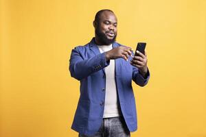 Shoppaholic african american man adding payment method on smartphone, studio background. Smiling BIPOC person does online shopping, typing credit card information on mobile phone photo