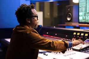 African american record producer adjusting sound settings on control board desk, editing tunes in professional studio. Music engineer working on mixing console to edit music, pushing faders. photo