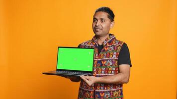 Tech content creator doing influencer marketing using green screen laptop, isolated over studio background. Indian man holding empty copy space mockup notebook, doing review, camera B photo