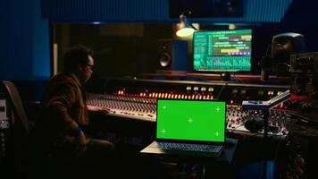 Music producer recording tracks next to laptop showing mockup isolated display, working with mixing console and mastering techniques in control room. Audio engineer producing new tunes. Camera B. photo