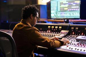 African american record composer creating new music for an album, editing and mixing tunes on console in control room. Audio technician using digital software on pc, adjust volume settings. photo