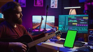Audio engineer practices tuning his acoustic guitar in home studio, watching a tutorial online via tablet with greenscreen. Musician learning to play instrument and create music. Camera B. photo
