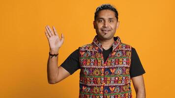 Portrait of jolly man smiling and waving hand, saluting, gesturing, isolated over studio backdrop. Joyous upbeat indian person in traditional attire doing greeting hand gesture, camera A photo